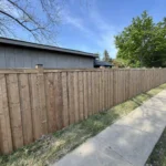 A wooden fence stands in front of a house, with a clear sidewalk leading up to the entrance.